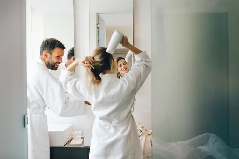 Man and woman standing together in front of bathroom mirror and smiling as the woman blow dries her hair