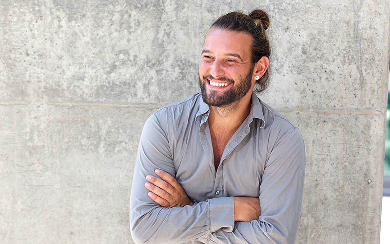 Man with a beard and a man bun leaning against a wall with his arms folded and smiling