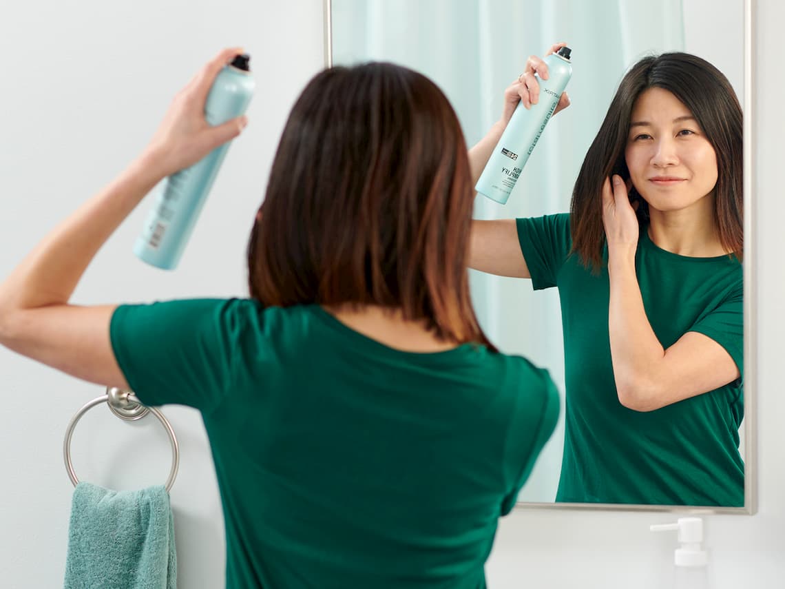 Woman looking in the mirror while styling her hair with hair spray
