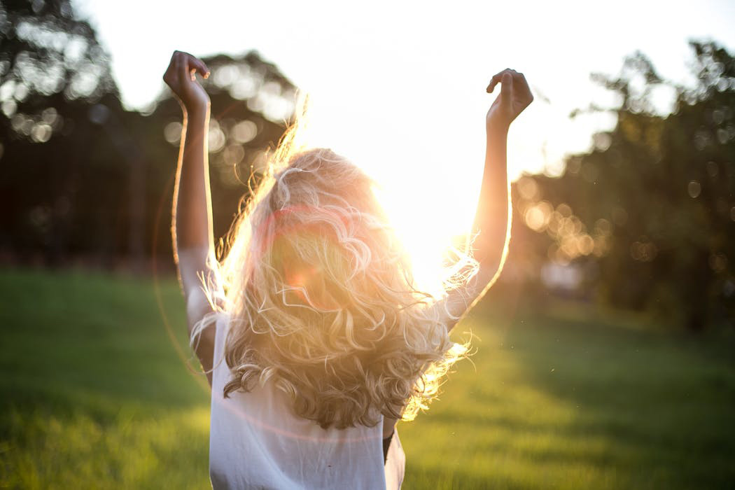 A woman with long curly hair standing with her back to us with her arms raised and the sun shining through her hair