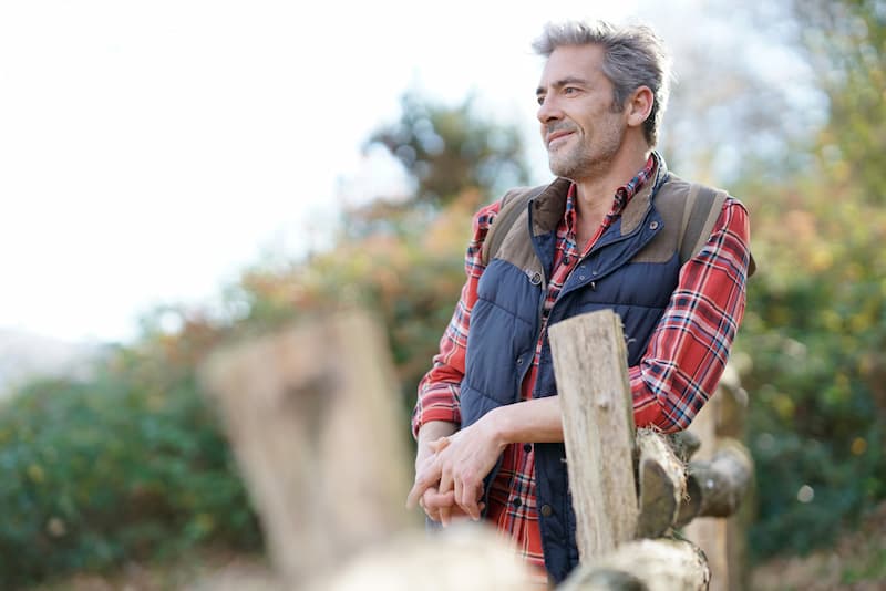 Man with gray hair leaning against a wooden fence smiling and staring into distance