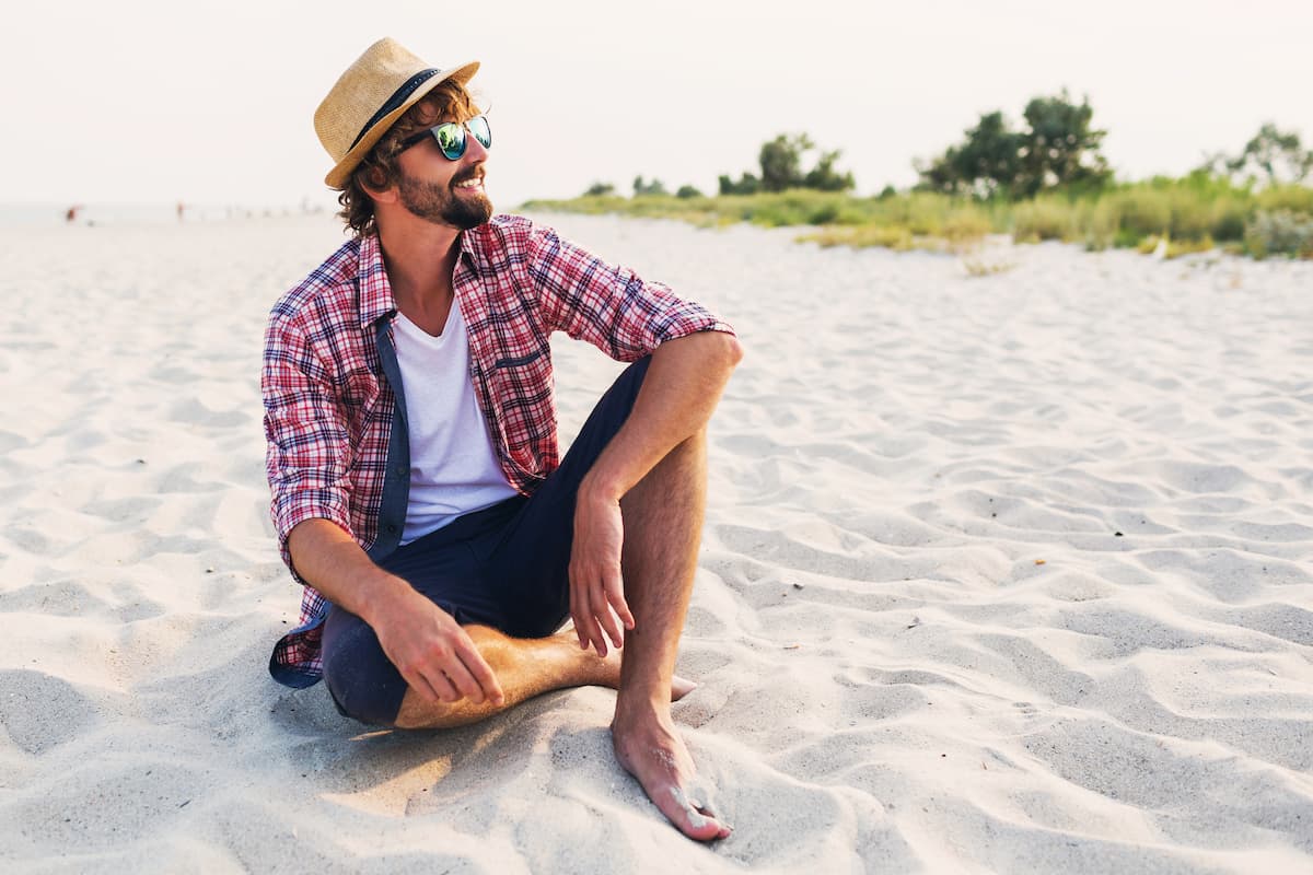 Young man wearing a hat and sunglasses sitting on the beach smiling