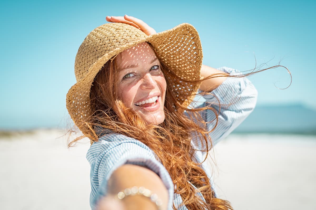 Woman with long, red, curly hair wearing a hat on a beach and taking a selfie